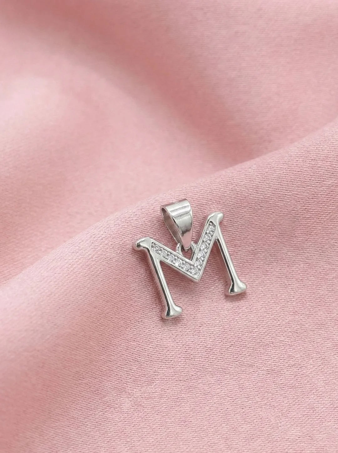 Silver letter 'M' pendant with clear stones on a pink fabric background