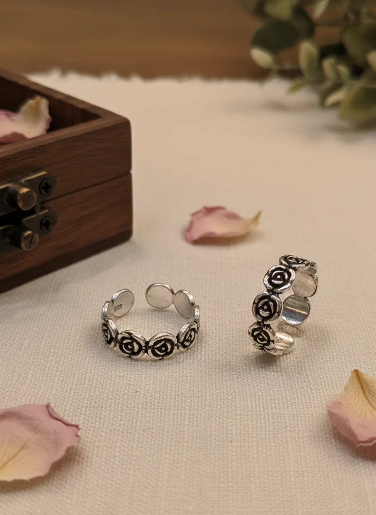 Silver toe rings with floral designs on a textured surface with pink petals and a wooden box.