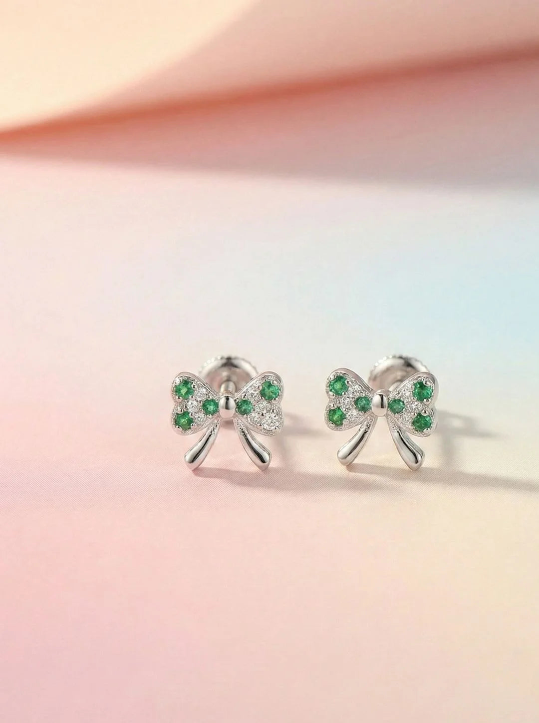 Bow-shaped earrings with green gemstones on a pastel pink and orange gradient background