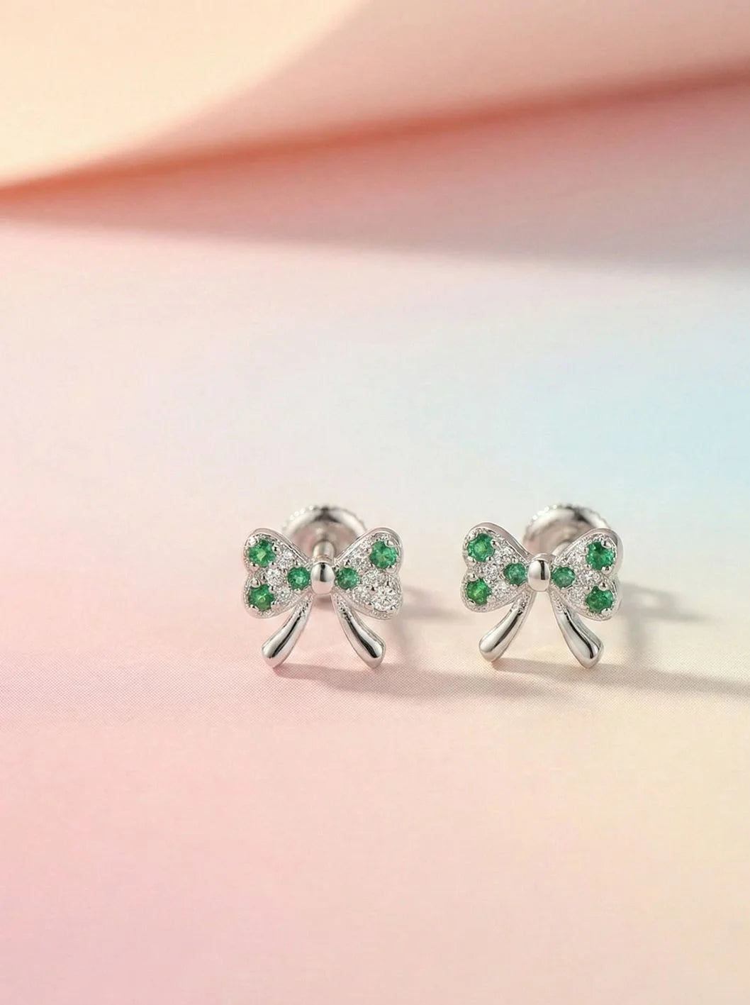 Bow-shaped earrings with green gemstones on a pastel pink and orange gradient background