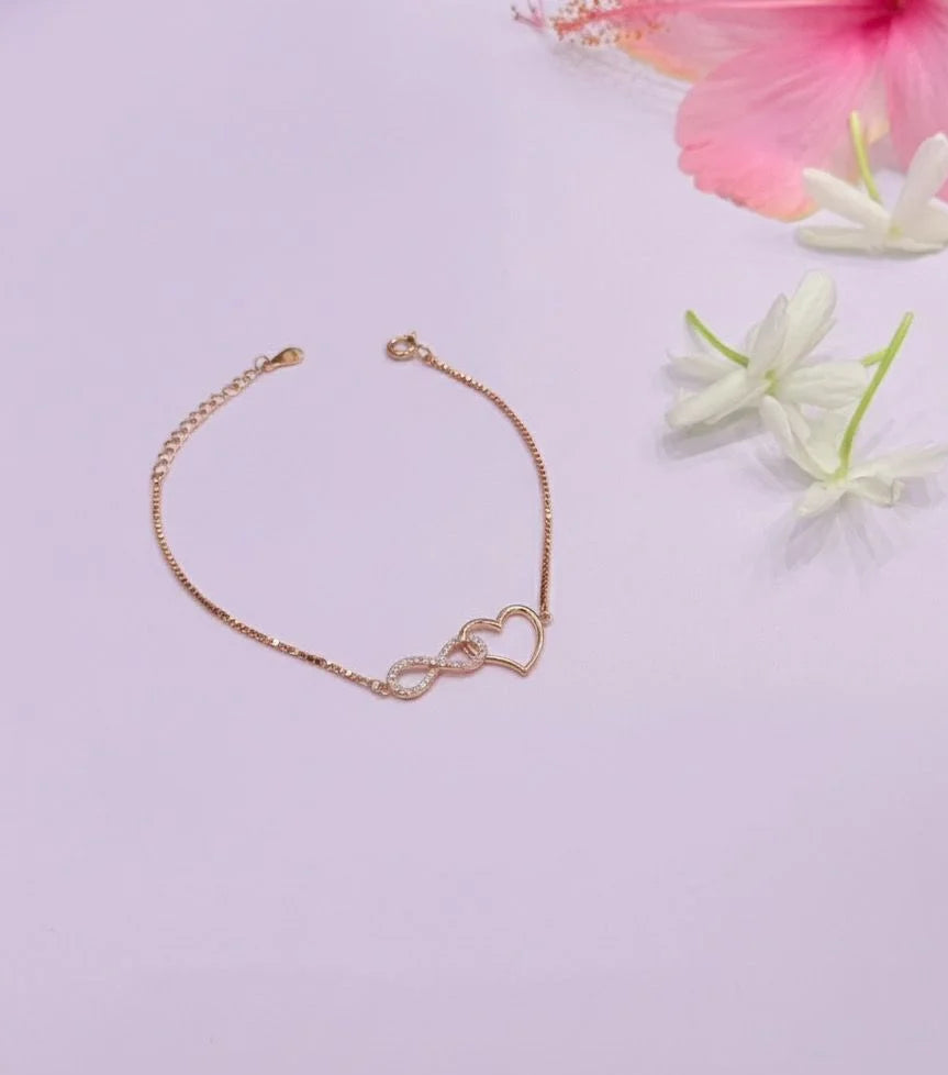 Rose gold bracelet with heart and infinity symbol on a light purple background with flowers.