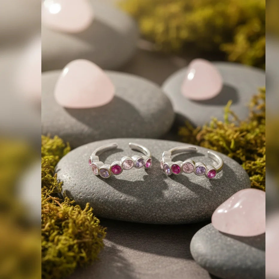 Silver toe rings with pink and purple stones on a stone surface with pink stones and greenery in the background