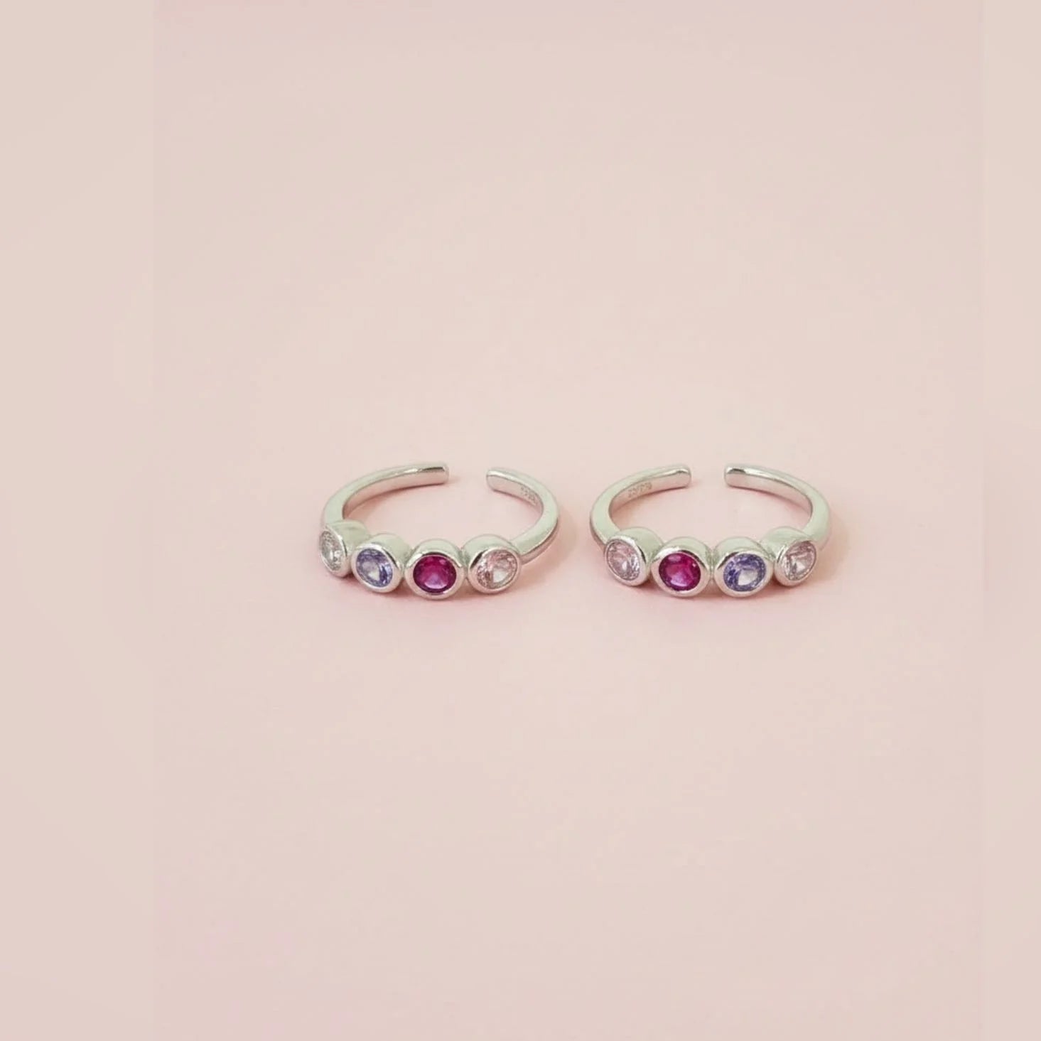 Two silver toe  rings with colorful gemstones on a pink background