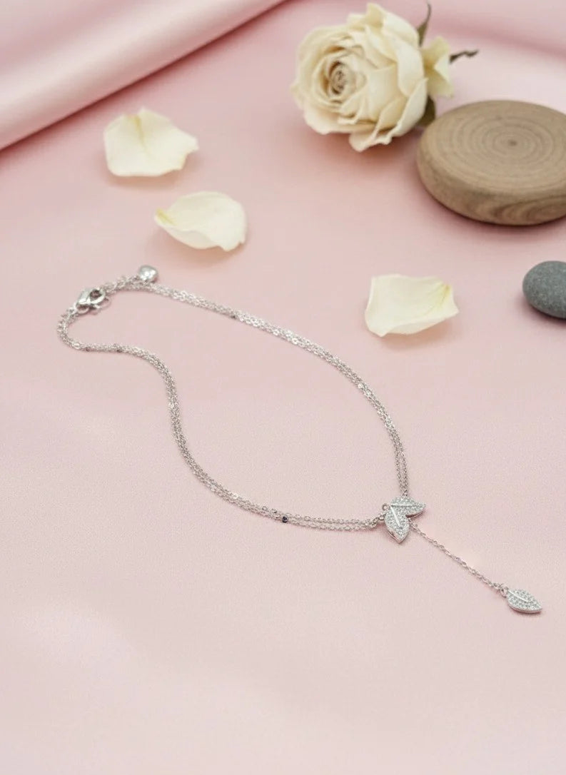 Silver necklace with a leaf pendant on a pink surface with flowers and stones.