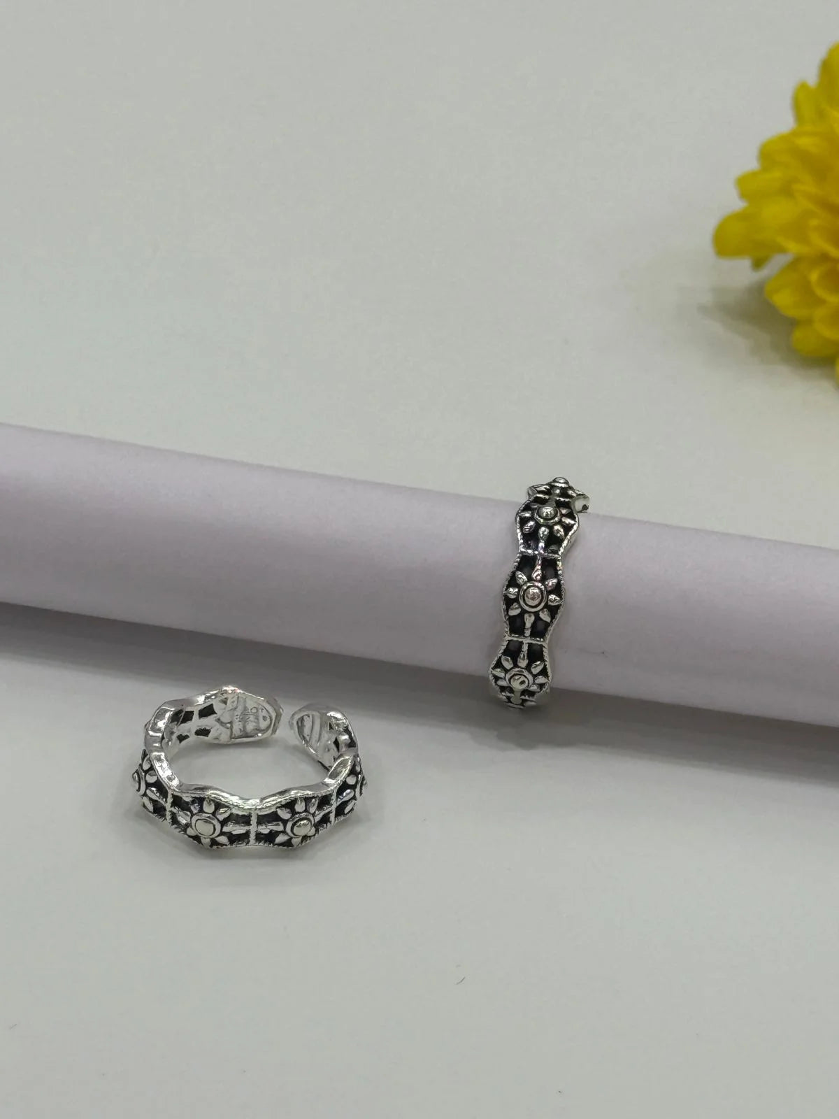 Two silver toe rings on a white surface with a yellow flower in the background