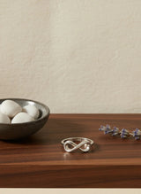 Silver infinity ring on a wooden surface with a bowl of white stones and dried lavender.