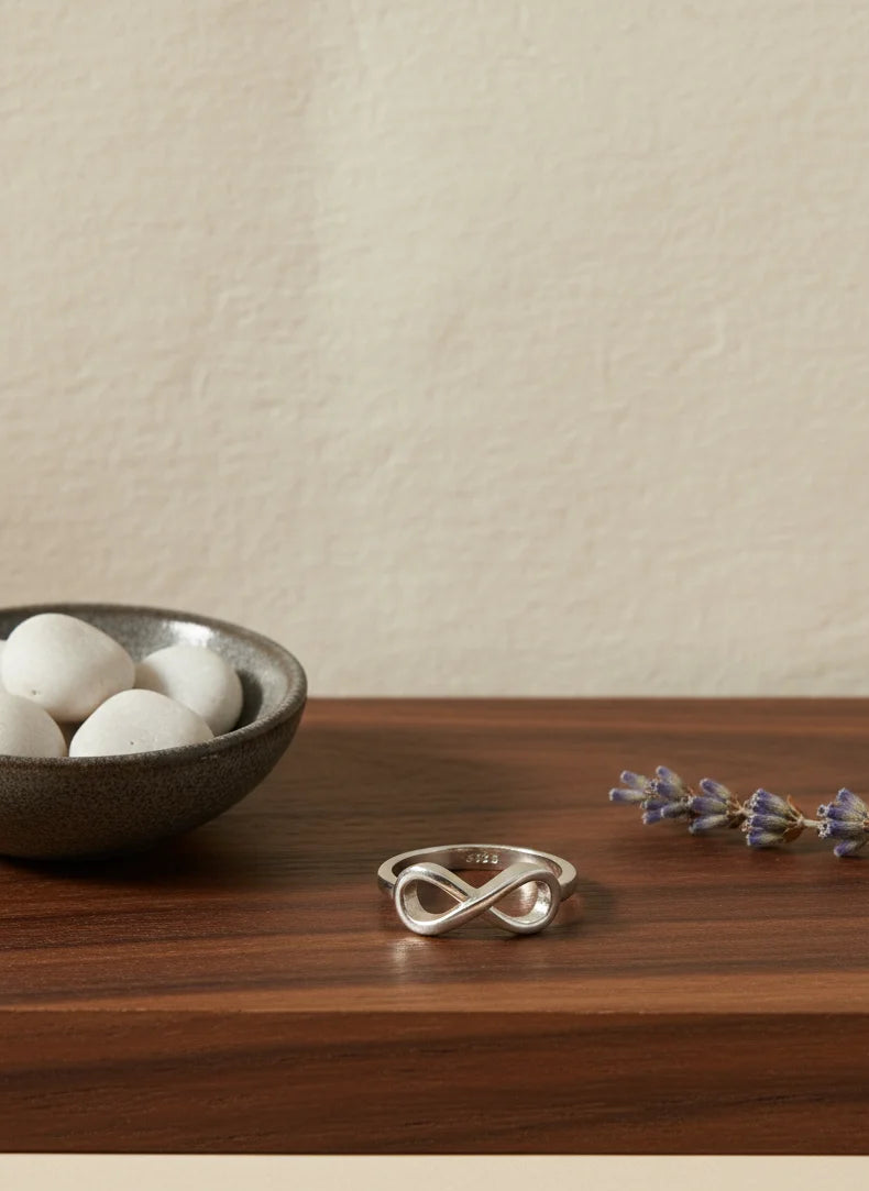 Silver infinity ring on a wooden surface with a bowl of white stones and dried lavender.