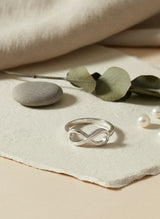Silver ring with infinity design on a beige surface with green leaves and pearls.