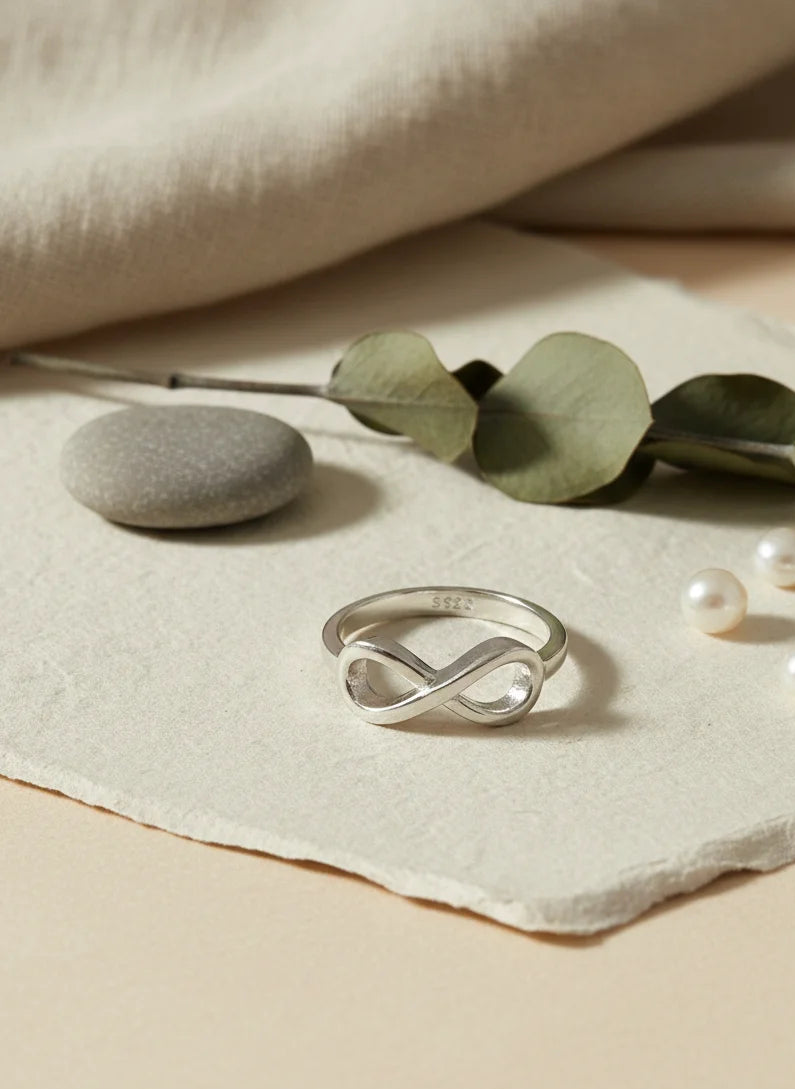 Silver ring with infinity design on a beige surface with green leaves and pearls.