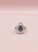 Teardrop-shaped gemstone ring with diamond halo on a pink background