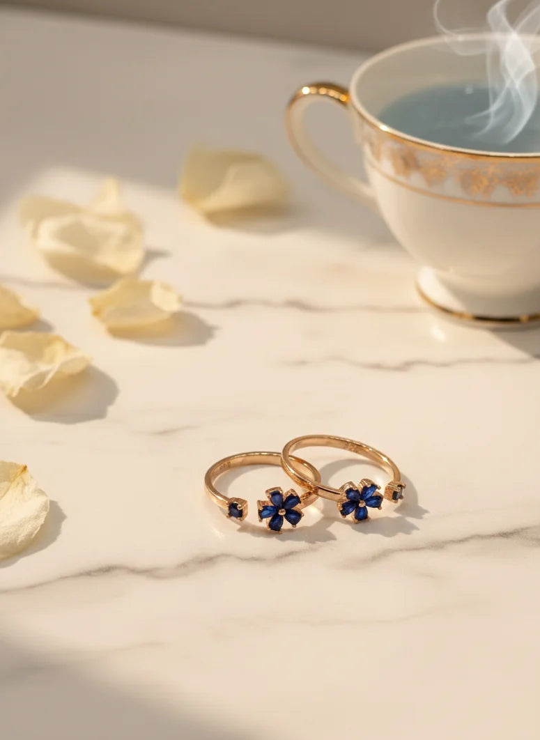 Two gold toe rings with blue stones on a marble surface next to a steaming cup of tea.