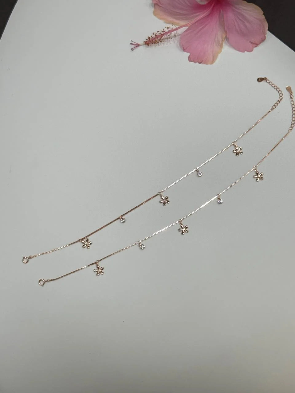 Silver anklet with floral pendants on a light gray background with a pink flower.