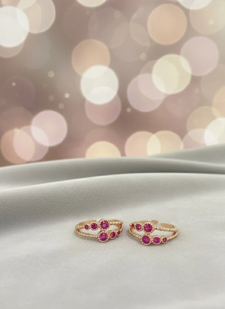Pair of gold toe rings with red gemstones on a white surface with a blurred bokeh background