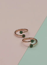 Two rose gold toe rings with green gemstones on a two-tone pink and green background