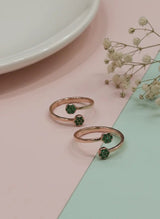 Two rose gold toe rings with green gemstones on a pastel pink and green background.