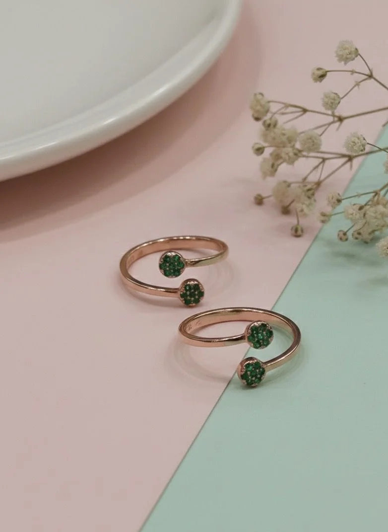 Two rose gold toe rings with green gemstones on a pastel pink and green background.