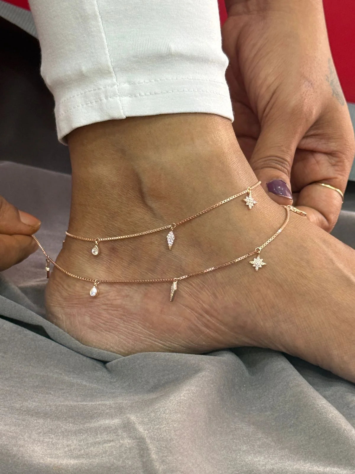 Close-up of a foot wearing multiple gold anklets with various charms on a neutral background.