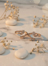 Two gold toe rings with diamond accents on a surface with white stones and dried plants.