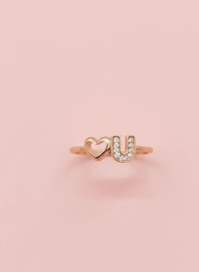 Rose gold ring with 'U' and heart design on a pink background