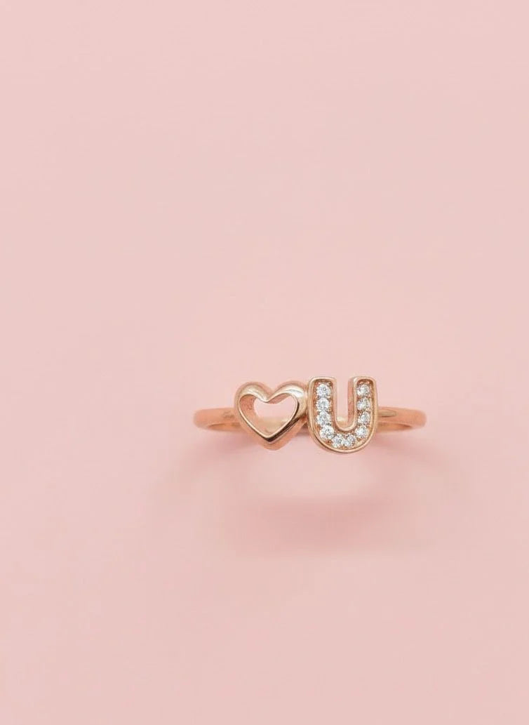 Rose gold ring with 'U' and heart design on a pink background