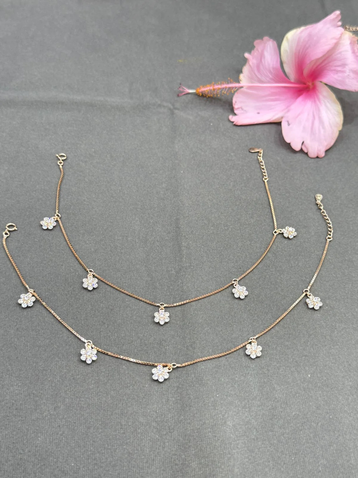 Two gold anklet  with floral pendants on a gray surface with a pink flower.