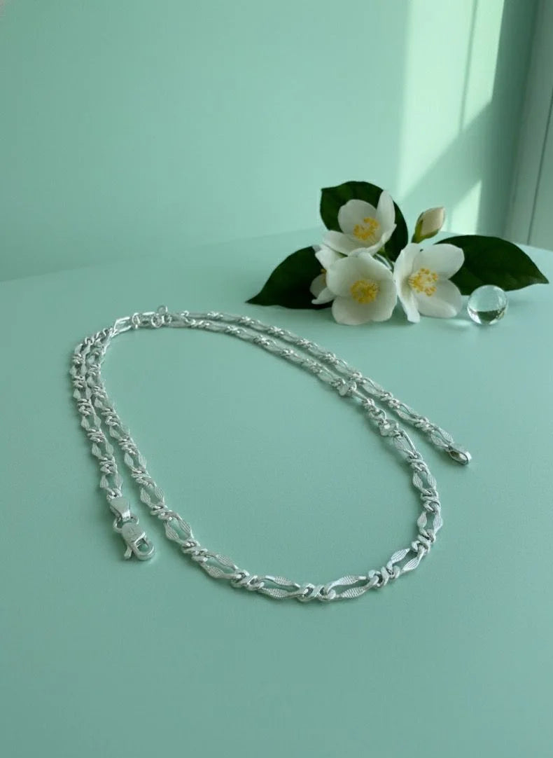 Silver chain necklace on a light green surface with white flowers and green leaves.
