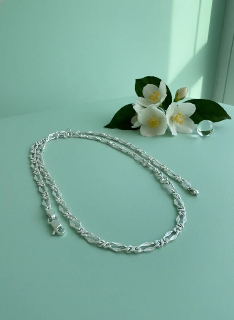 Silver chain necklace on a light green surface with white flowers and green leaves.