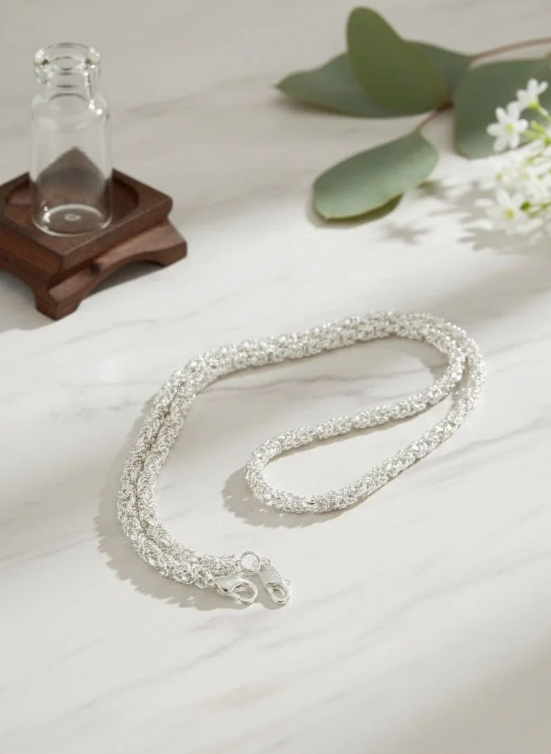 Silver necklace on a light surface with a glass bottle and greenery in the background
