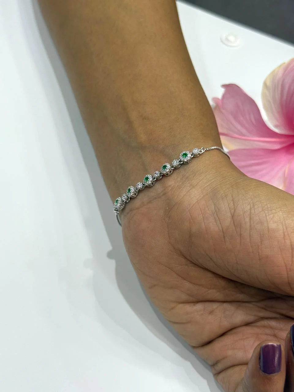 Hand wearing a silver bracelet with green stones on a white surface with pink petals.