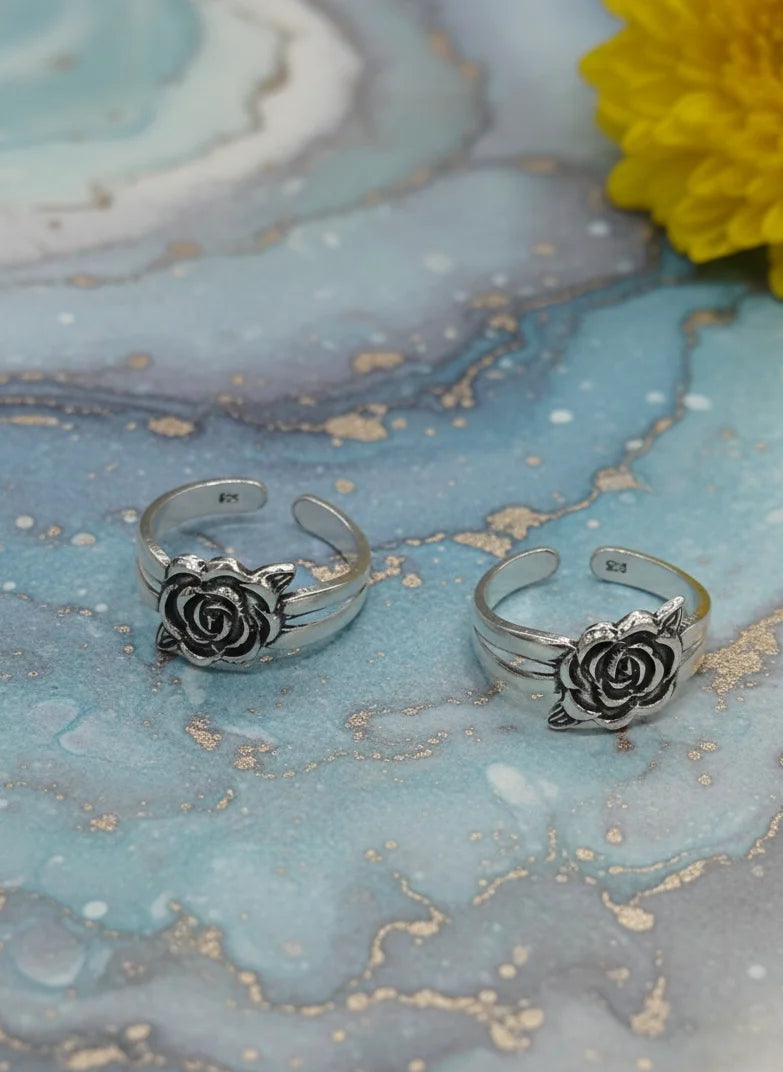 Silver rose-shaped toe rings on a marble surface with a yellow flower.