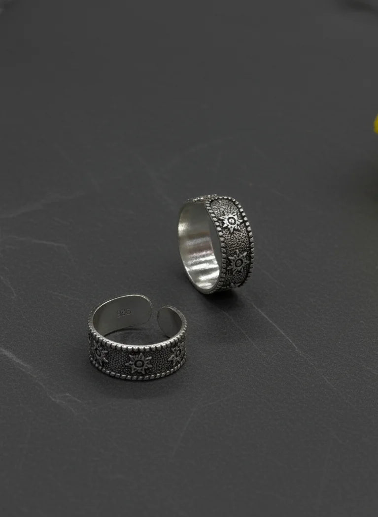 Two silver toe rings with intricate designs on a dark textured surface with a yellow flower.