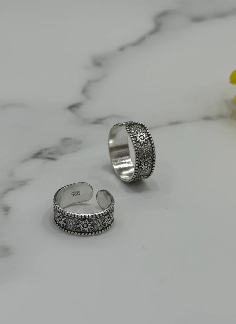 Two silver toe rings with floral designs on a marble surface with a yellow flower.