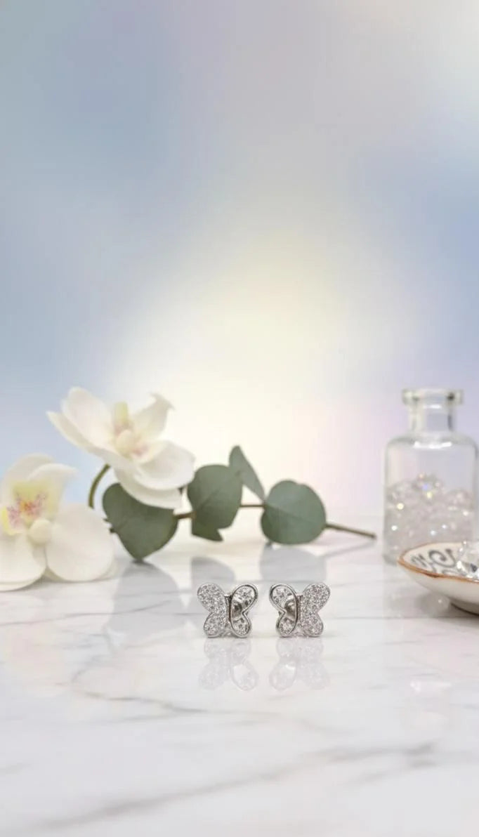 Silver butterfly earrings on a white surface with flowers and a bottle in the background