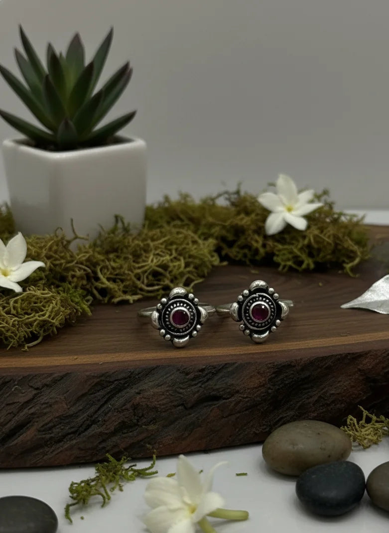 Two silver toe rings with red stones on a wooden surface with flowers and rocks.
